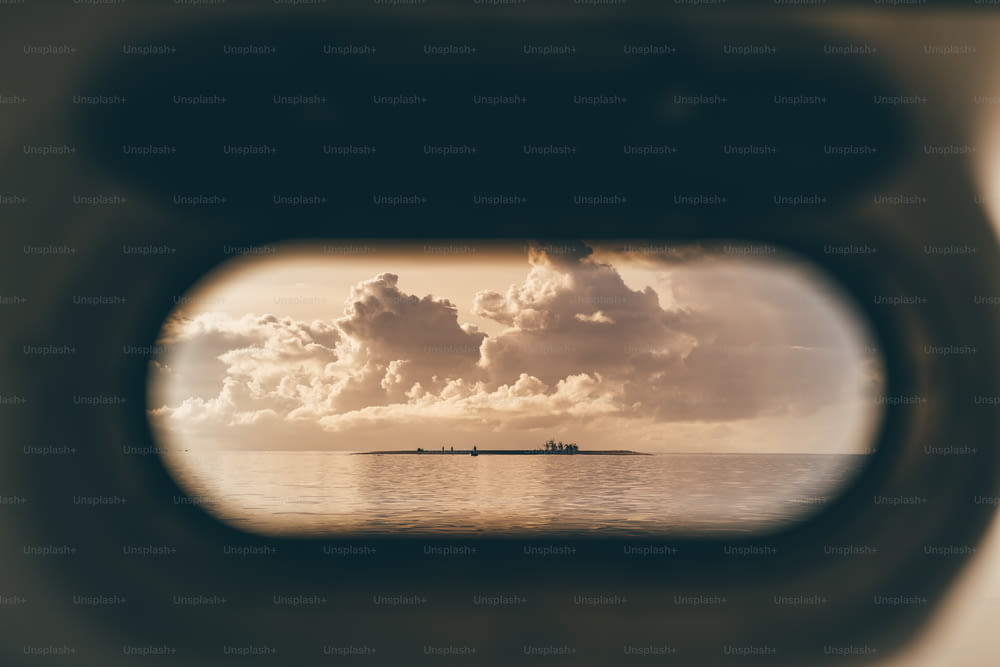 Vue à travers le hublot ouvert d’un navire d’une petite île entourée par l’eau de l’océan le soir, avec des silhouettes de personnes dessus et un magnifique paysage de nuages au coucher du soleil au-dessus, Maldives