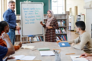 Mujer musulmana con hiyab de pie en la pizarra haciendo una tarea con verbos irregulares durante la lección de inglés