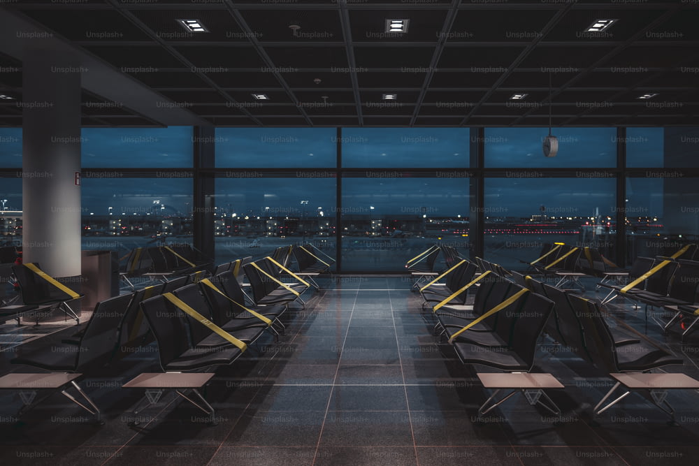 Una veduta grandangolare di una sala d'attesa buia, vuota e abbandonata in quarantena di un moderno terminal aeroportuale di notte, in lockdown con regolari nastri verdastri sui sedili per mantenere il distanziamento sociale