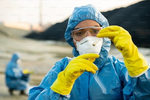 Young researcher in protective workwear holding sample of toxic soil in flask in front of her face during ecological investigation with colleague
