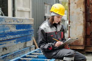 Young serious female engineer in protective helmet and workwear making sketch on paper in clipboard while sitting on building materials