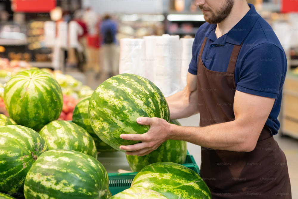 Unrecognizable supermarket worker wearing uniform setting out fresh watermelons, copy space