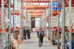 Young mixed-race warehouse worker in uniform moving along aisle between large racks against two colleagues scrolling through data