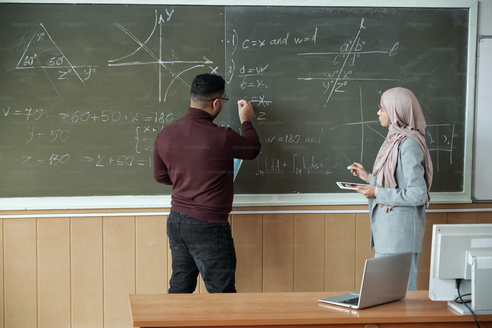 ヒジャーブをかぶった男性教授と若い女子学生が黒板のそばに立って授業で方程式を解き、男性が導関数の1つを指差す