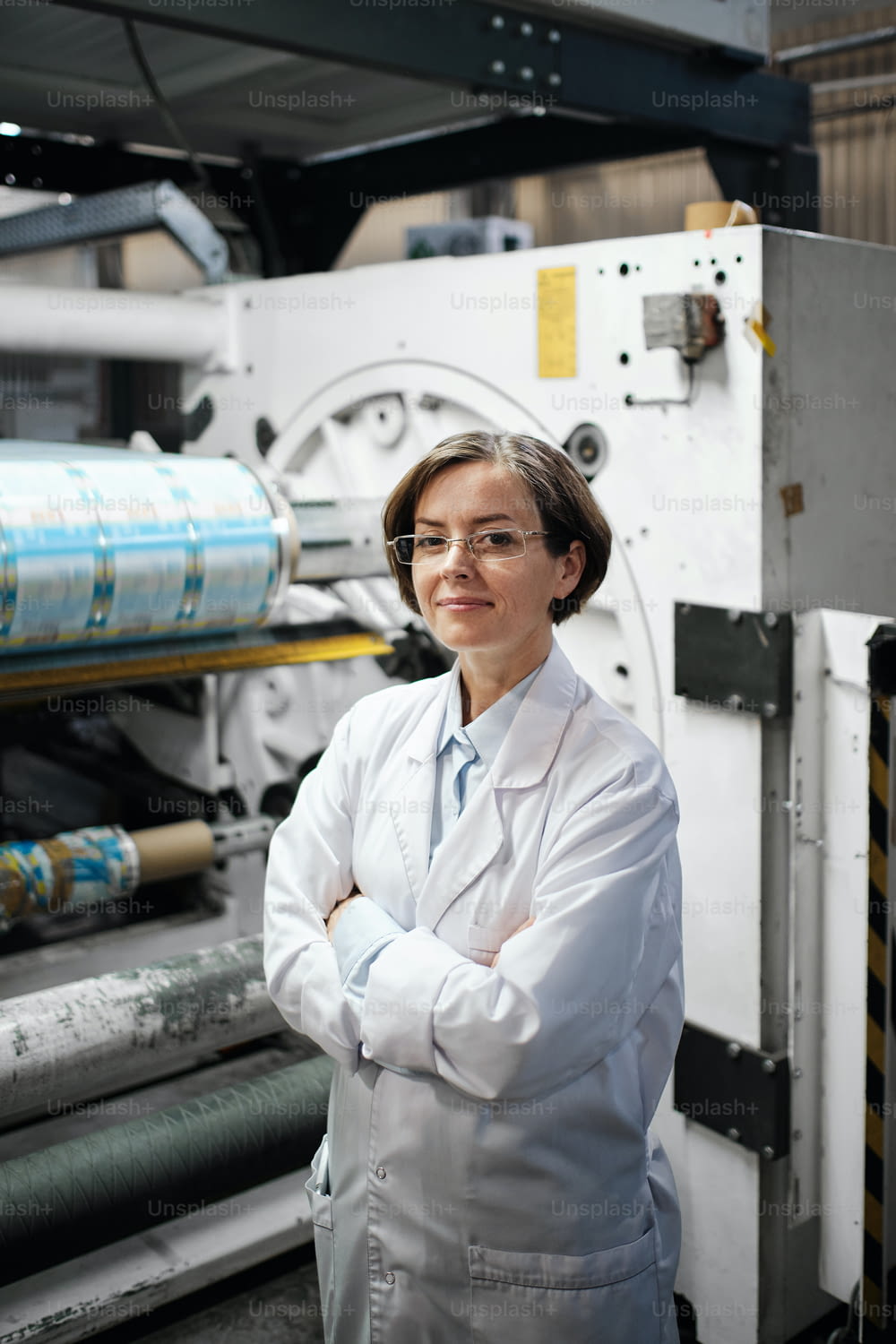 Portrait of female factory worker wearing glasses and white robe at polymer plastic manufacturing, doing quality control and checking machines and mechanisms