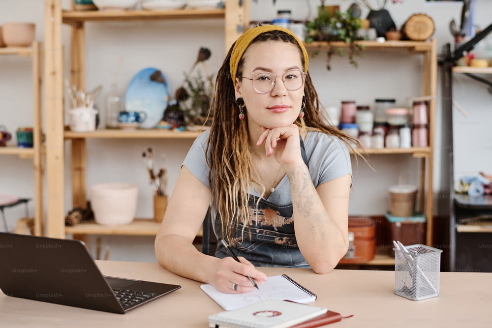 Young modern female in casualwear and eyeglasses sitting by workplace in studio and looking at camera while making sketch in notepad