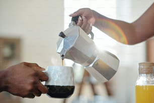Hand of young African female pouring coffee or tea into mug of her husband during breakfast with bottle of orange juice near by