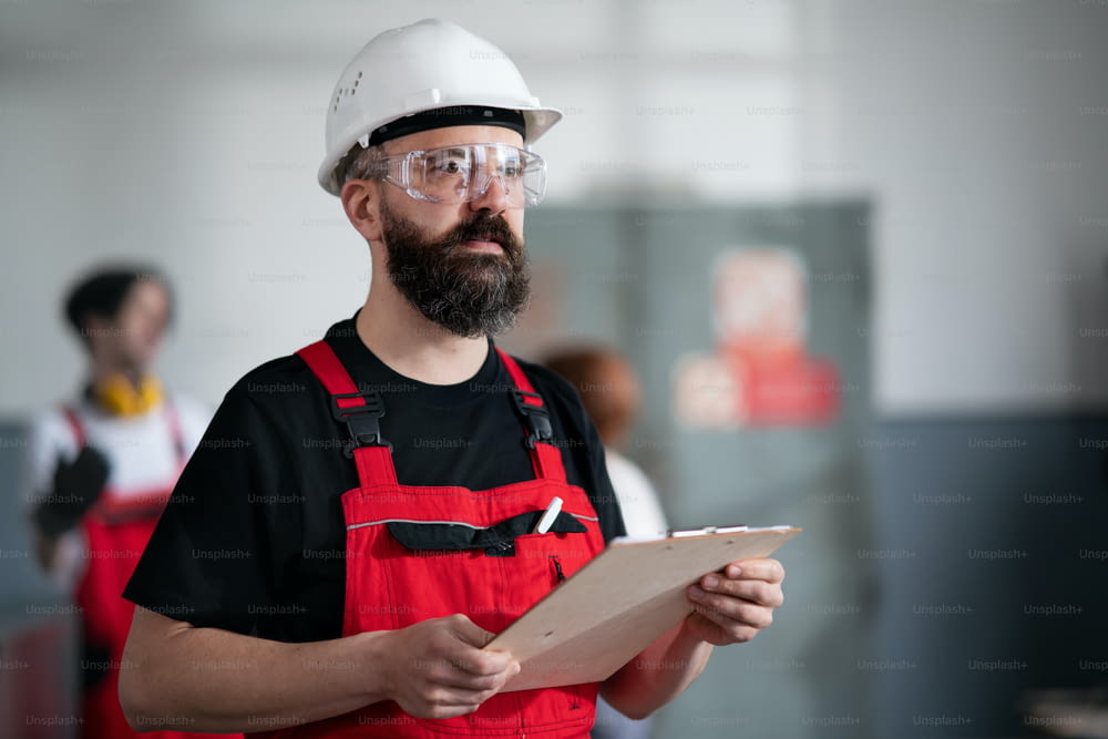 A portrait of worker with helmet and protective glasses indoors in factory holding clipboard.