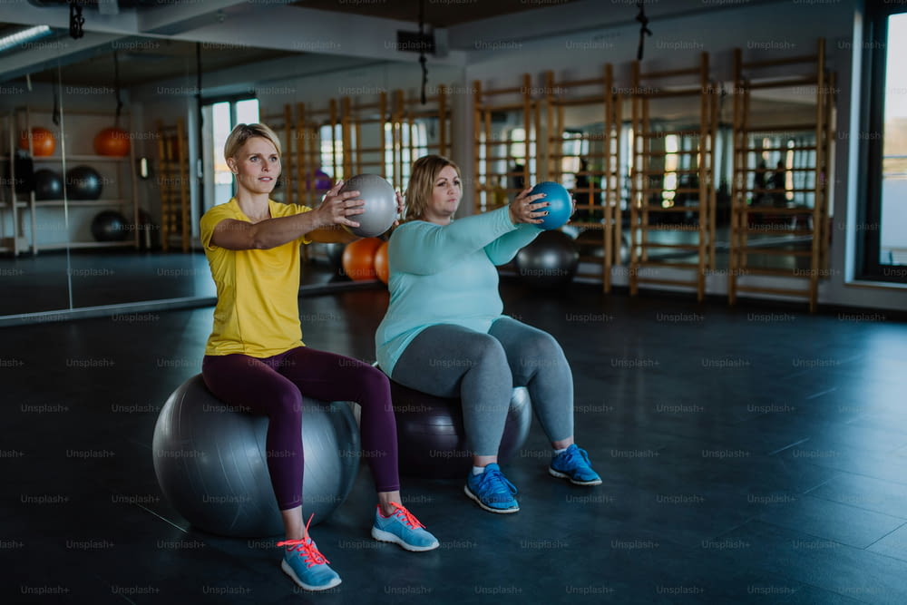 An overweight woman with dumbbells sitting on fintess ball and exercising with personal trainer in gym
