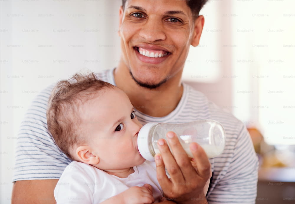 A portrait of cheerful father bottle feeding a small toddler son indoors at home.