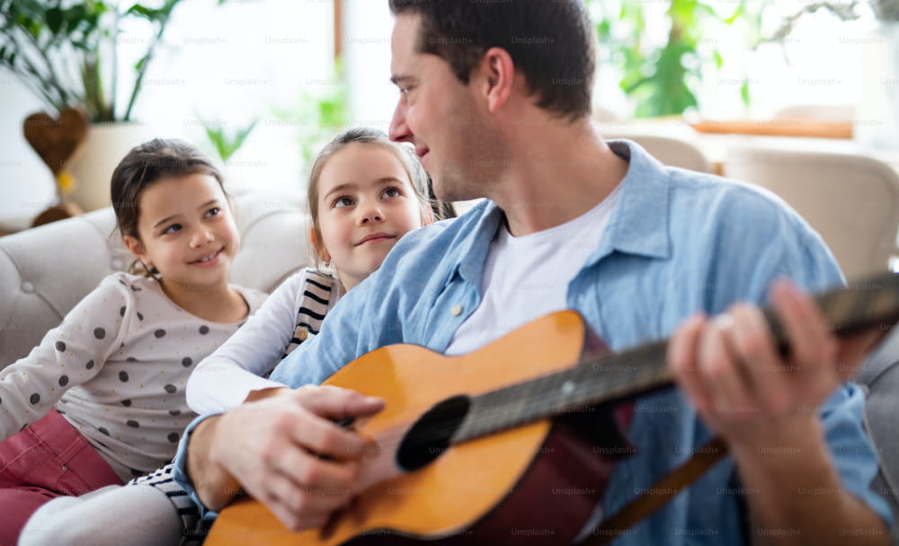 Portrait of father with small daughters sitting on sofa indoors at home, playing guitar.