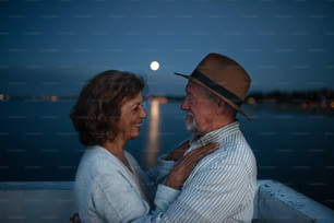 A happy senior couple hugging outdoors on pier by sea at dusk, looking at each other.