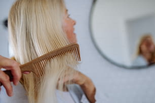 A beautiful senior woman in bathrobe combing hair with wooden comb in bathroom, sustainable lifestyle.