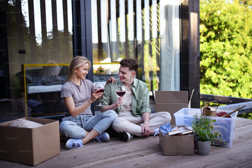 A cheerful young couple moving in their new tiny house in woods sitting on floor on terrace and drinking wine. Conception of moving and sustainable living.