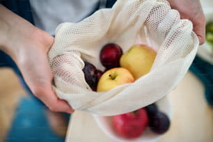 Midsection of woman holding fruit in reusable bag indoors at home, sustainable lifestyle concept.
