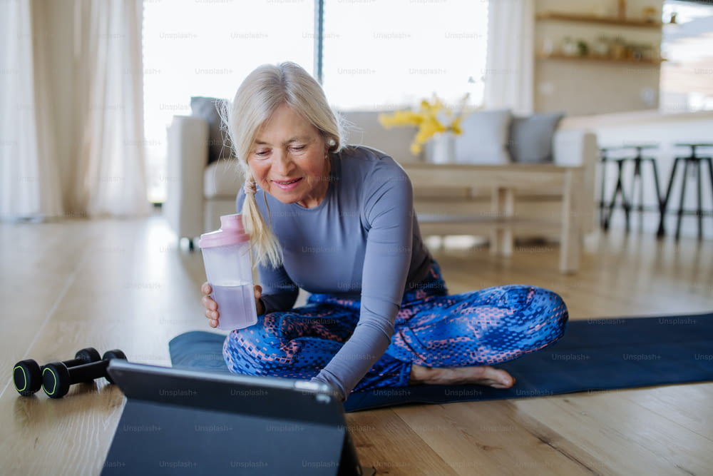 A fit senior woman doing stretching exercise at home with tutorial on tablet, active lifestyle concept.