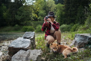 Front view of young woman with a dog on a walk outdoors in summer nature, taking photographs.