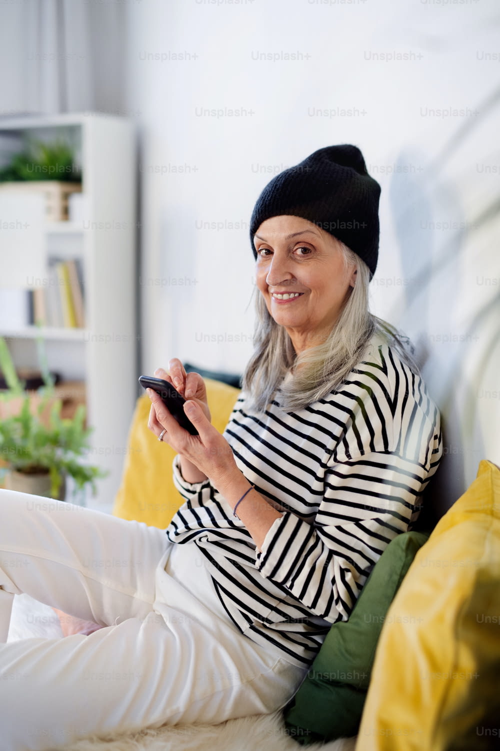 집에서 실내에 앉아 스마트폰을 사용하는 모자를 쓴 노인 여성의 초상화.