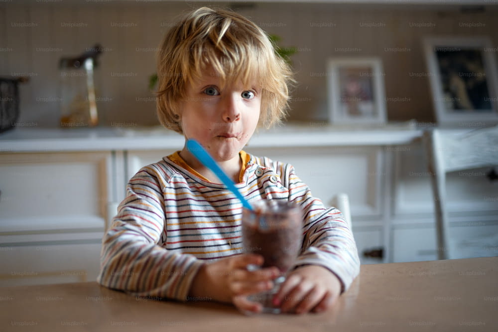 Portrait d’un petit garçon à la bouche sale à l’intérieur dans la cuisine à la maison, mangeant du pudding.