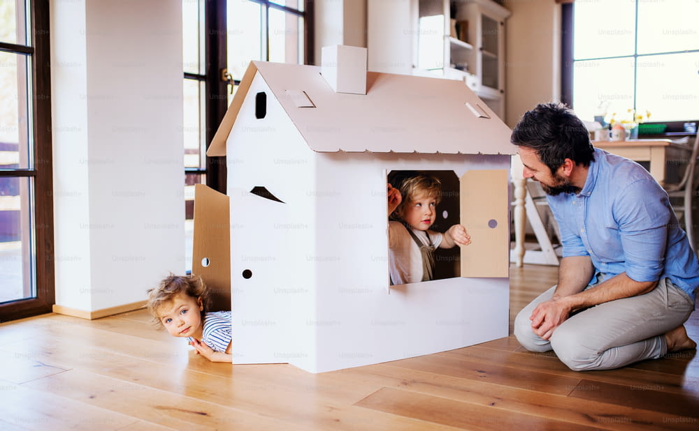 Two happy toddler children with father playing with paper house indoors at home.