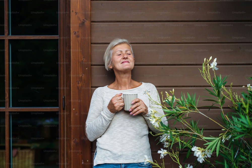 A senior woman with coffee standing outdoors on a terrace in summer, eyes closed.