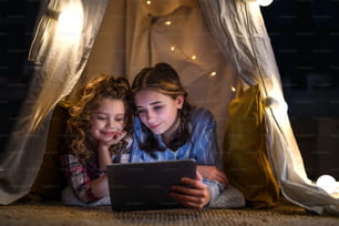 Sisters indoors at home, using tablet in pajamas in tent. Lockdown concept.