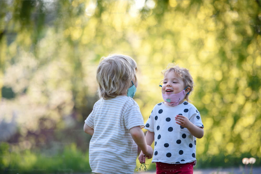 Small children with face masks playing outdoors in countryside, coronavirus concept.