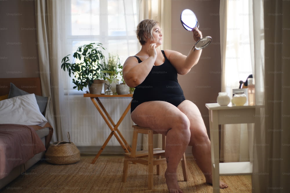 Una mujer con sobrepeso sentada y mirándose en el espejo en casa, concepto de cuidado personal.