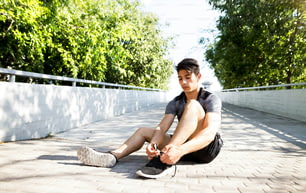 Young hispanic runner in the city sitting on concrete path tying his shoelaces.