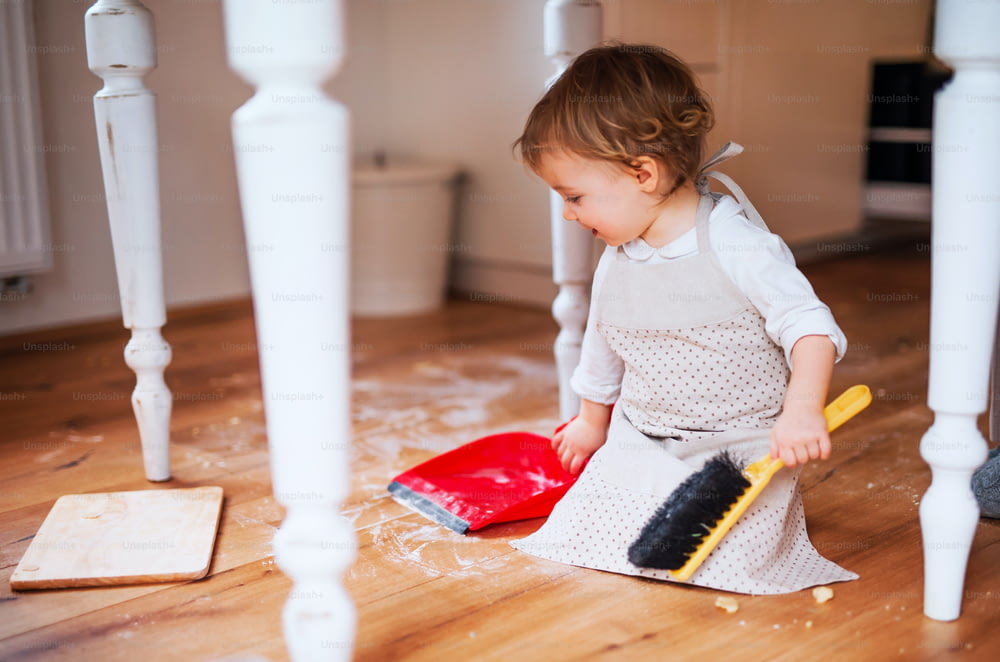 Une petite fille en bas âge avec une brosse et une pelle à poussière balayant le sol en désordre dans la cuisine à la maison.
