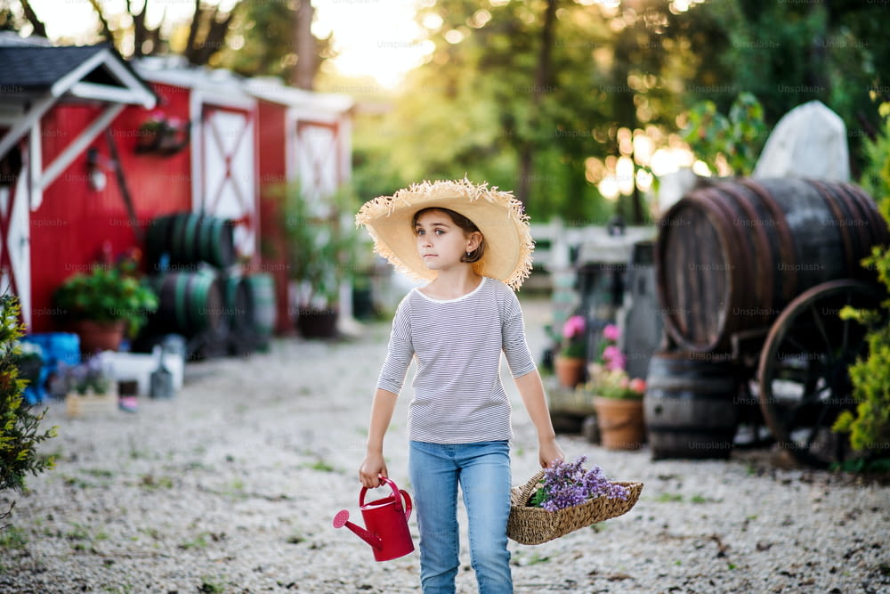A front view of small girl with a hat walking outdoors on family farm, holding plants.