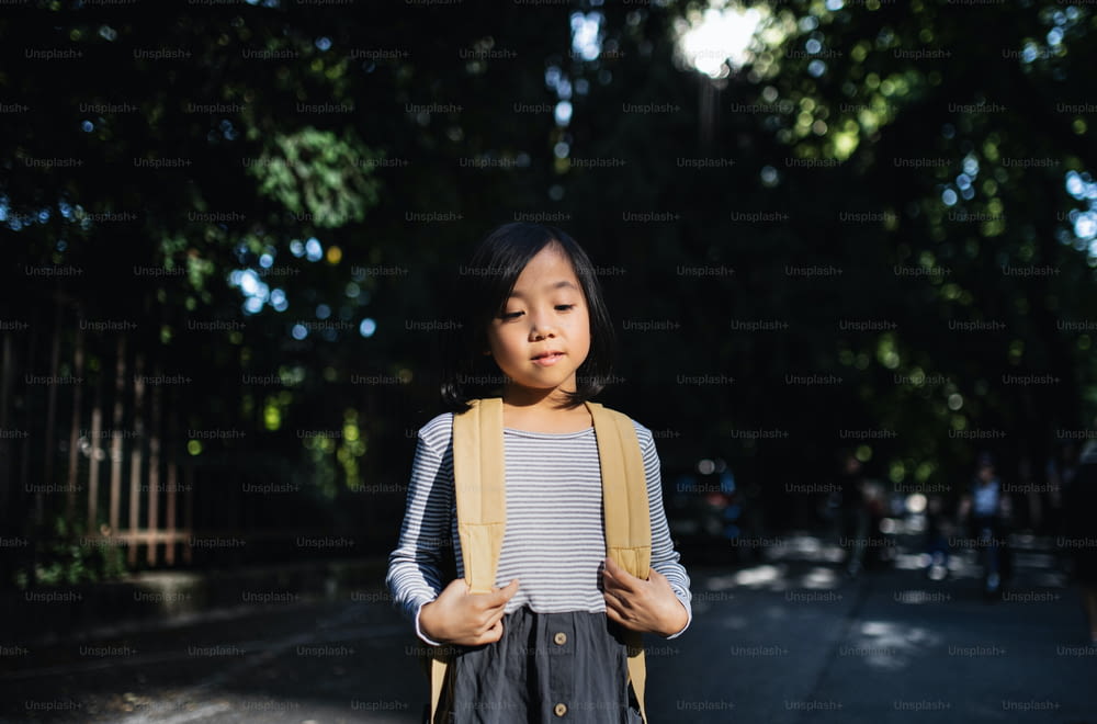 Front view portrait of small Japanese girl with backpack standing outdoors in town.