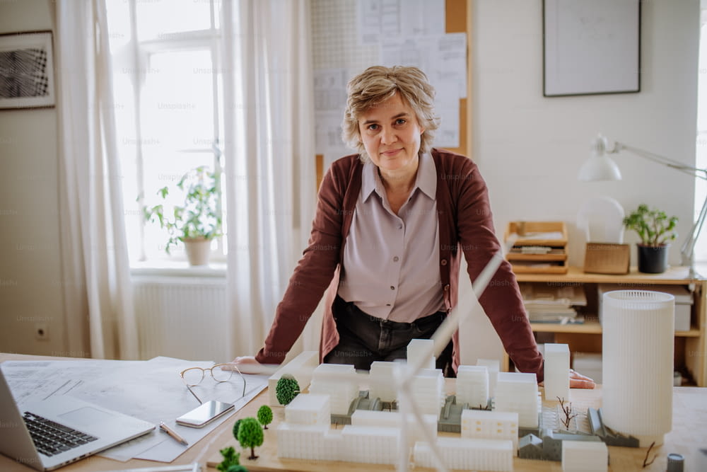 Happy senior woman architect with model of houses standing in office and looking at camera.