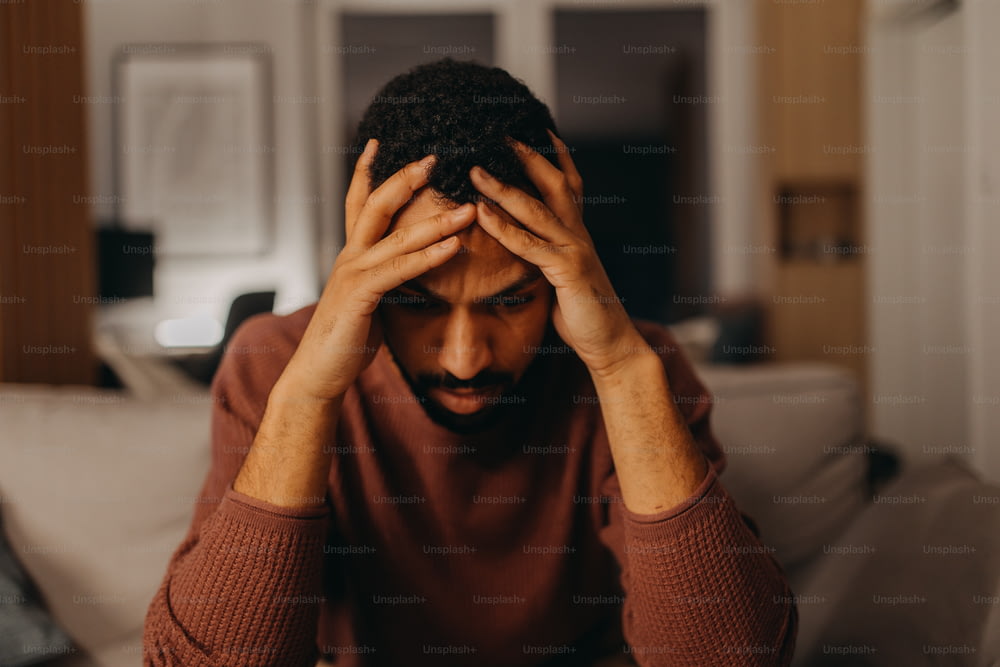 Um retrato od deprimido jovem afro-americano homem com a cabeça nas mãos sentado em casa.