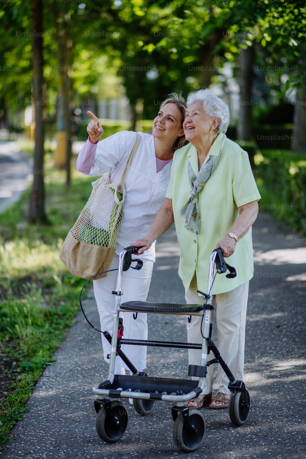 A caregiver with senior woman on walk with walker in park with shopping bag.