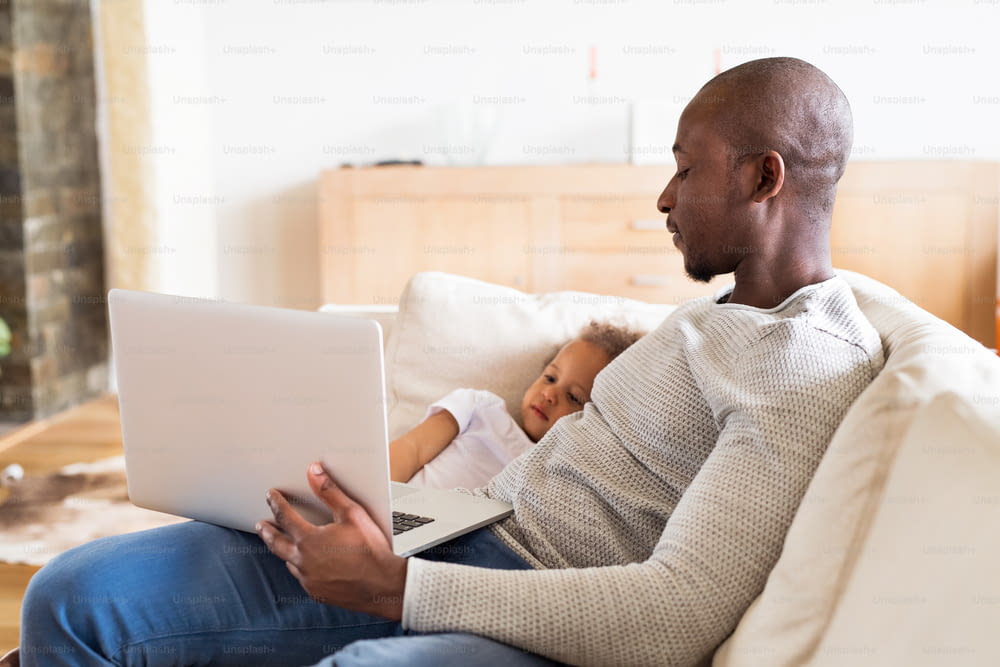 Joven padre afroamericano en casa con su linda hijita sentada en el sofá viendo algo en la computadora portátil.