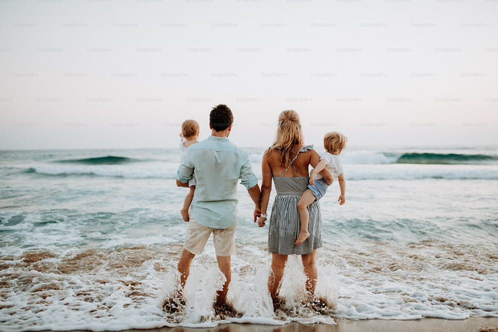 Eine Rückansicht der jungen Familie mit zwei Kleinkindern, die im Sommerurlaub am Strand stehen.