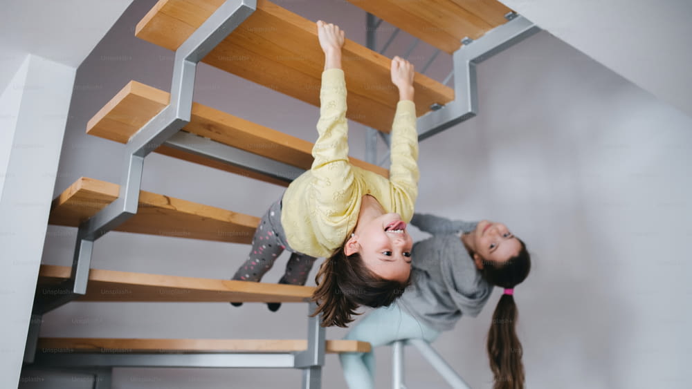 Two cheerful small girls sisters indoors at home, having fun under staircase.