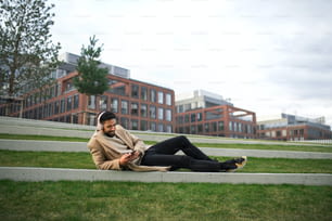 A man with headphones and smartphone outdoors in park in city, resting.