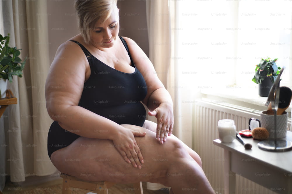 Uma mulher gorda sentada e aplicando creme nas pernas em casa, conceito de autocuidado.