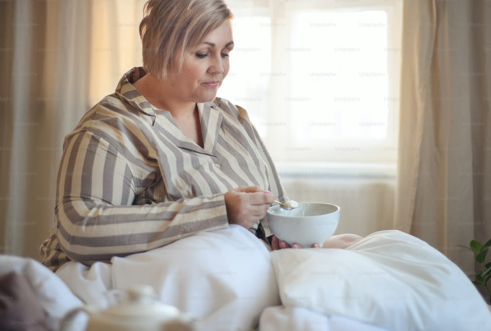 A happy overweight woman with headphones and smartphone having breakfast in bed at home.