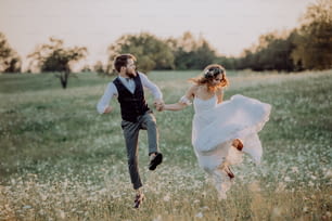 Belle jeune mariée et le marié à l’extérieur dans la nature verdoyante au coucher du soleil romantique, se tenant la main, sautant haut.