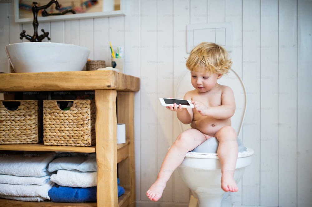 バスルームでスマートフォンを持つかわいい幼児。トイレに座っている小さな男の子。