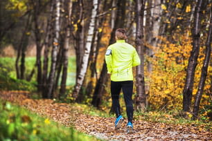 Joven atleta con teléfono inteligente en chaqueta amarilla corriendo afuera. Trail runner entrenando para correr a campo traviesa en la colorida y soleada naturaleza otoñal.
