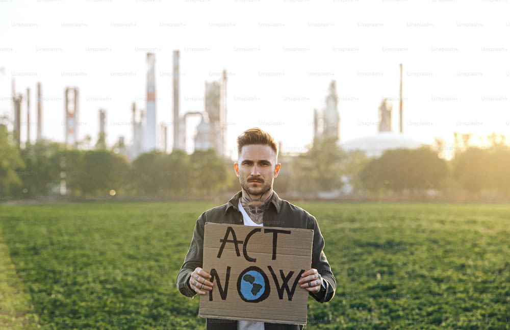 Retrato de un joven activista con un cartel de pie al aire libre junto a una refinería de petróleo, protestando.
