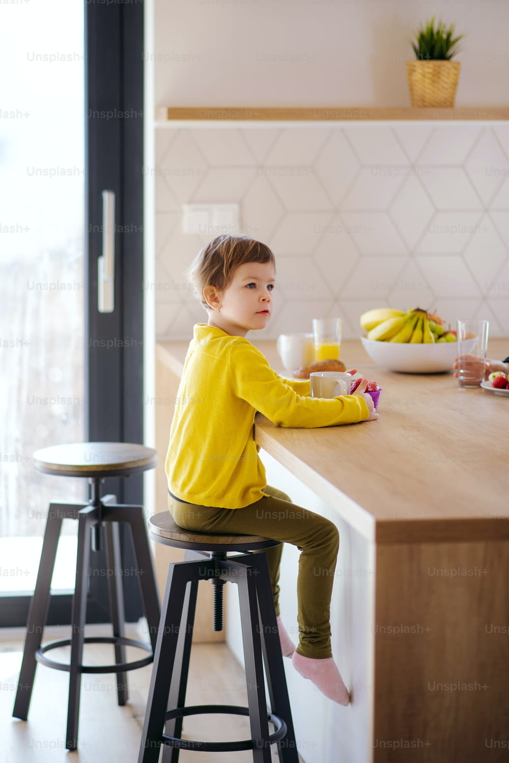 Una niña pequeña sentada en un taburete en la cocina de su casa, comiendo fruta.