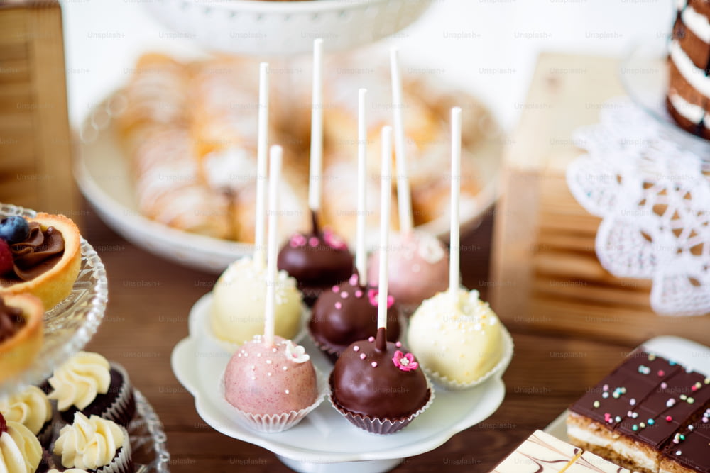 Table with white, pink and chocolate cake pops on a plate, tarts and cupcakes on cakestand. Candy bar.