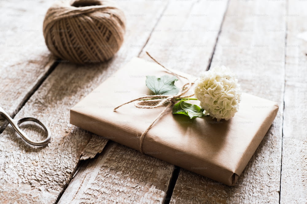 Cadeau enveloppé dans du papier brun décoré d’une fleur de lilas. Des ciseaux et une pelote de laine posés sur la table. Prise de vue en studio sur fond en bois blanc.