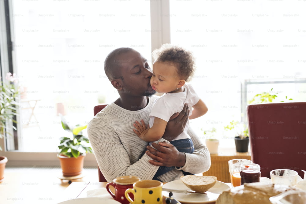 Young afro-american father at home with his little daughter holding her in arms, kissing her on cheek, having breakfast together.