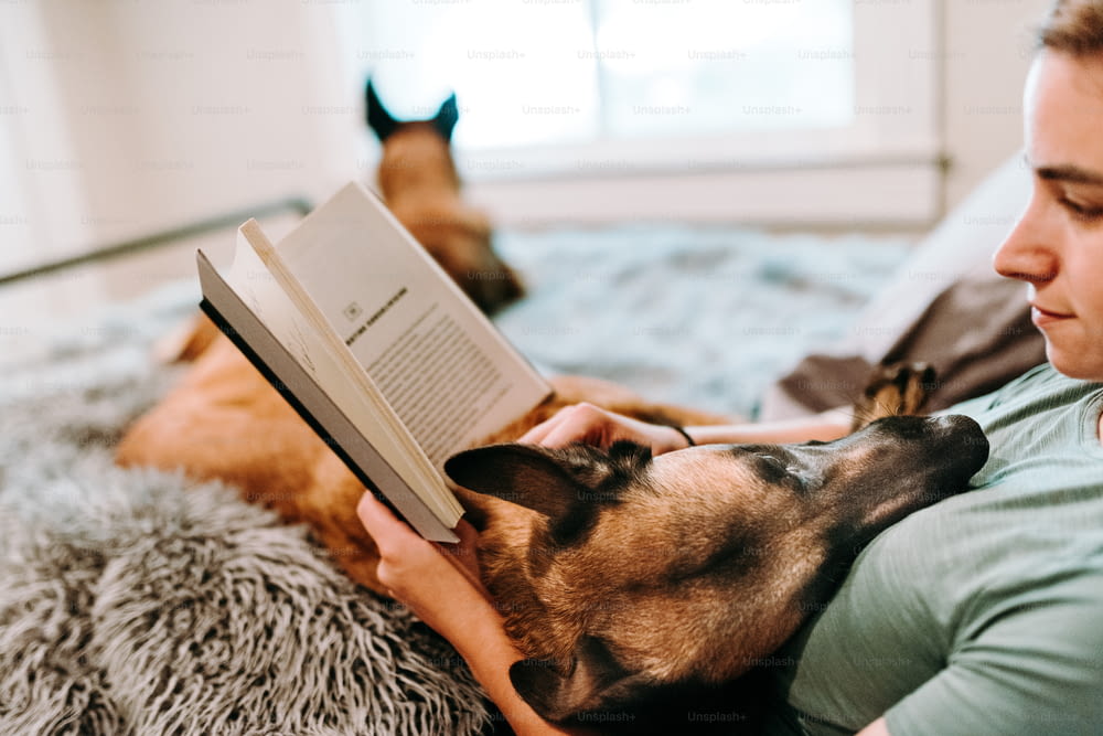 a woman laying on a bed reading a book to a dog